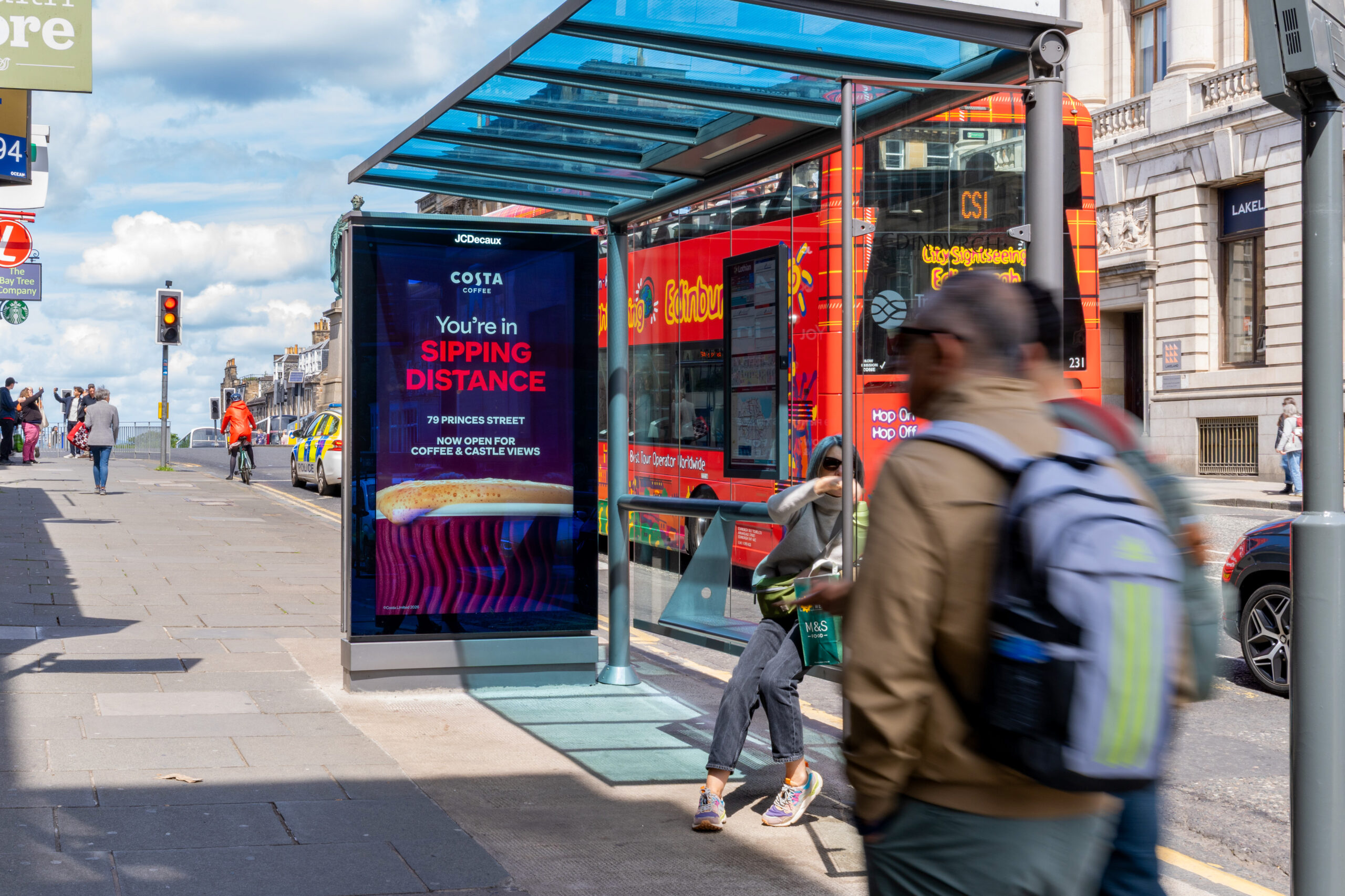Bus stop shelter with a blue digital ad reading 'You're in SIPPING DISTANCE' beside a red double-decker bus on a city street.