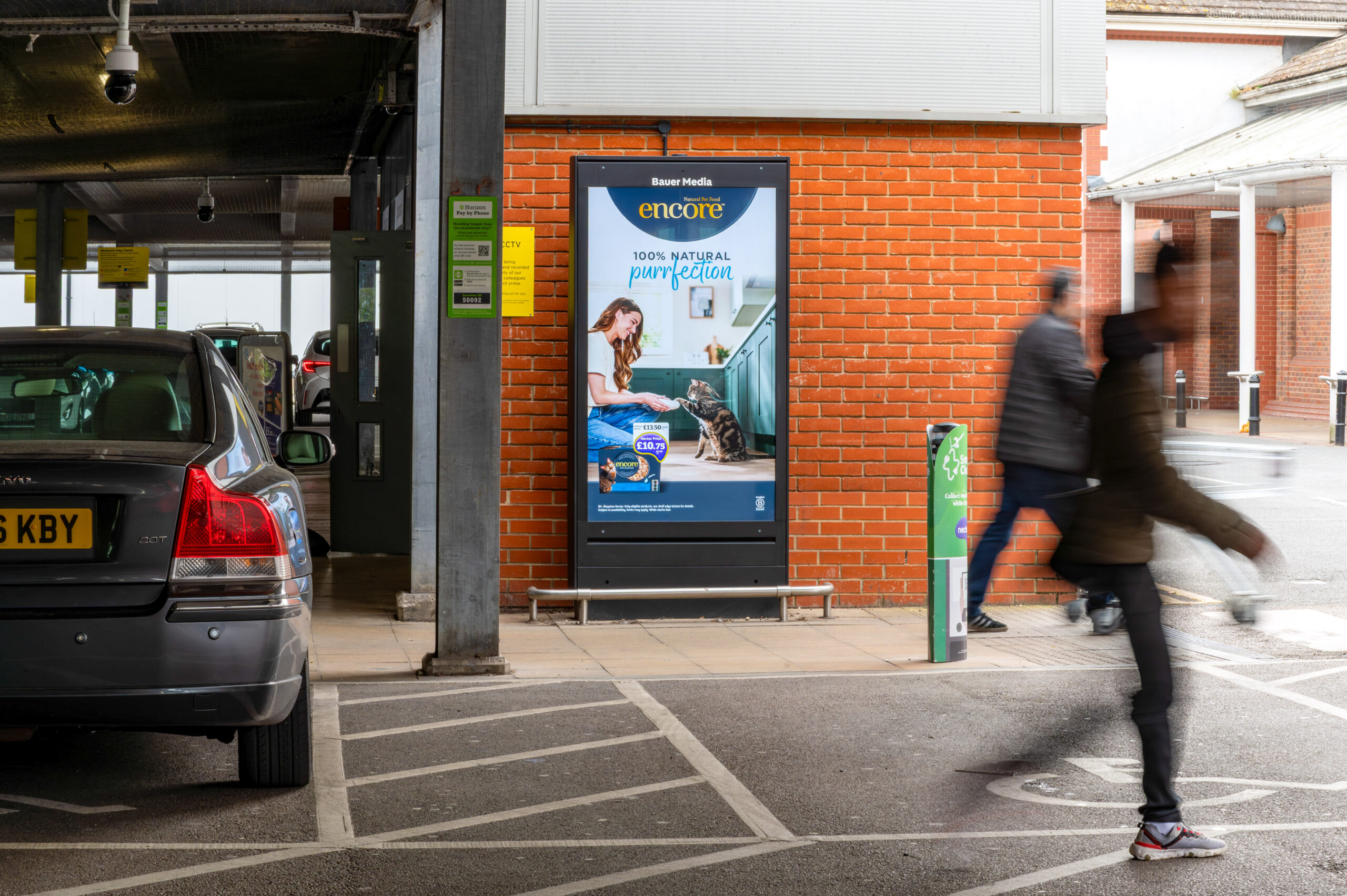 Digital ad board on a red brick wall beside a parking area, with blurred pedestrians walking by.