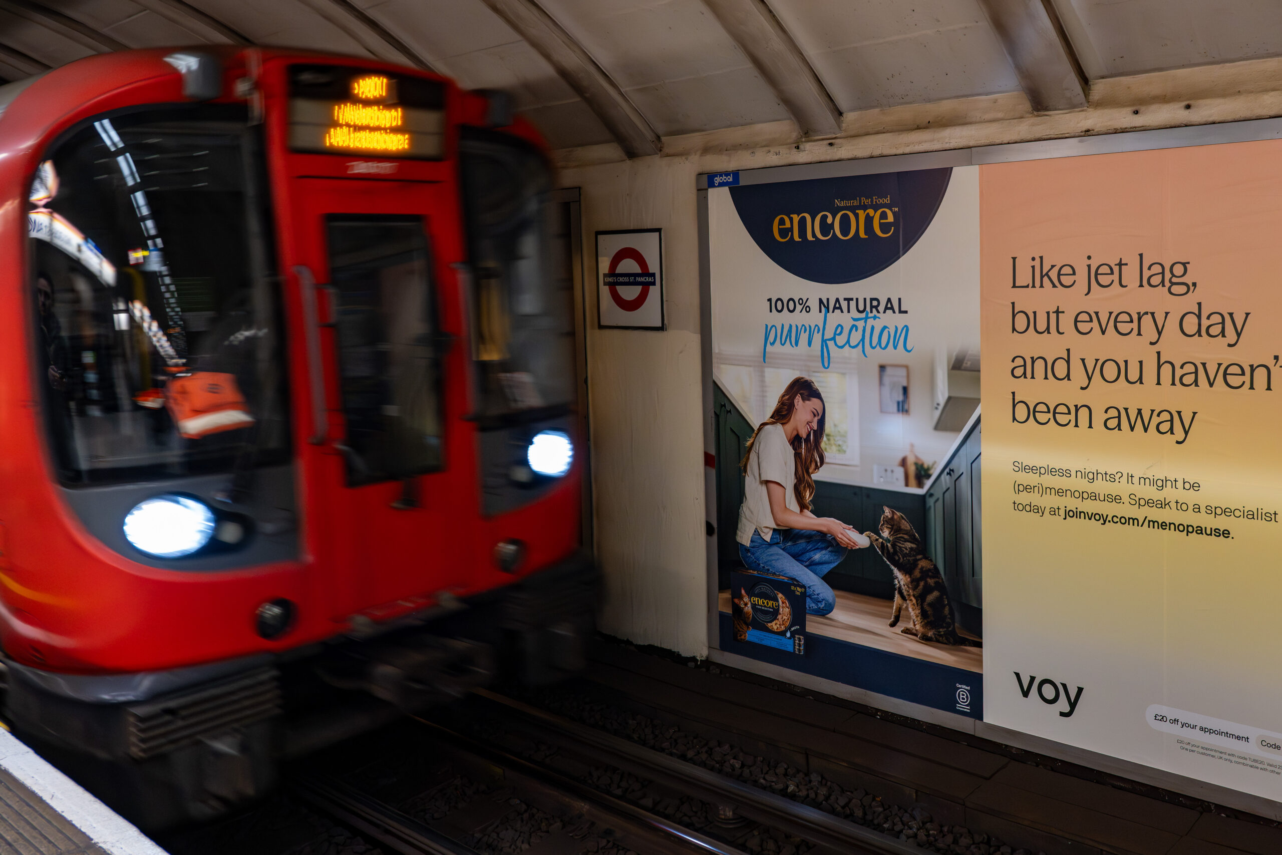 Red subway train rushing past a platform with large wall ads featuring 'encore' and a woman on the right side.