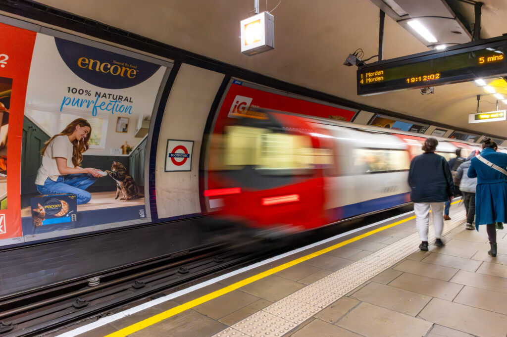 Subway platform as a red train speeds by; posters advertising Encore dog food on the wall, passengers waiting nearby.