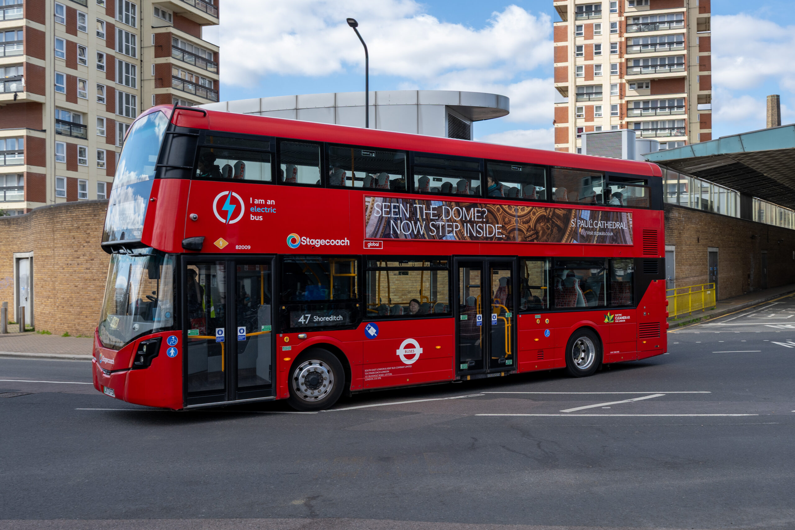 Red double-decker bus with advertisement on the side, driving past tall apartment buildings under a blue sky.