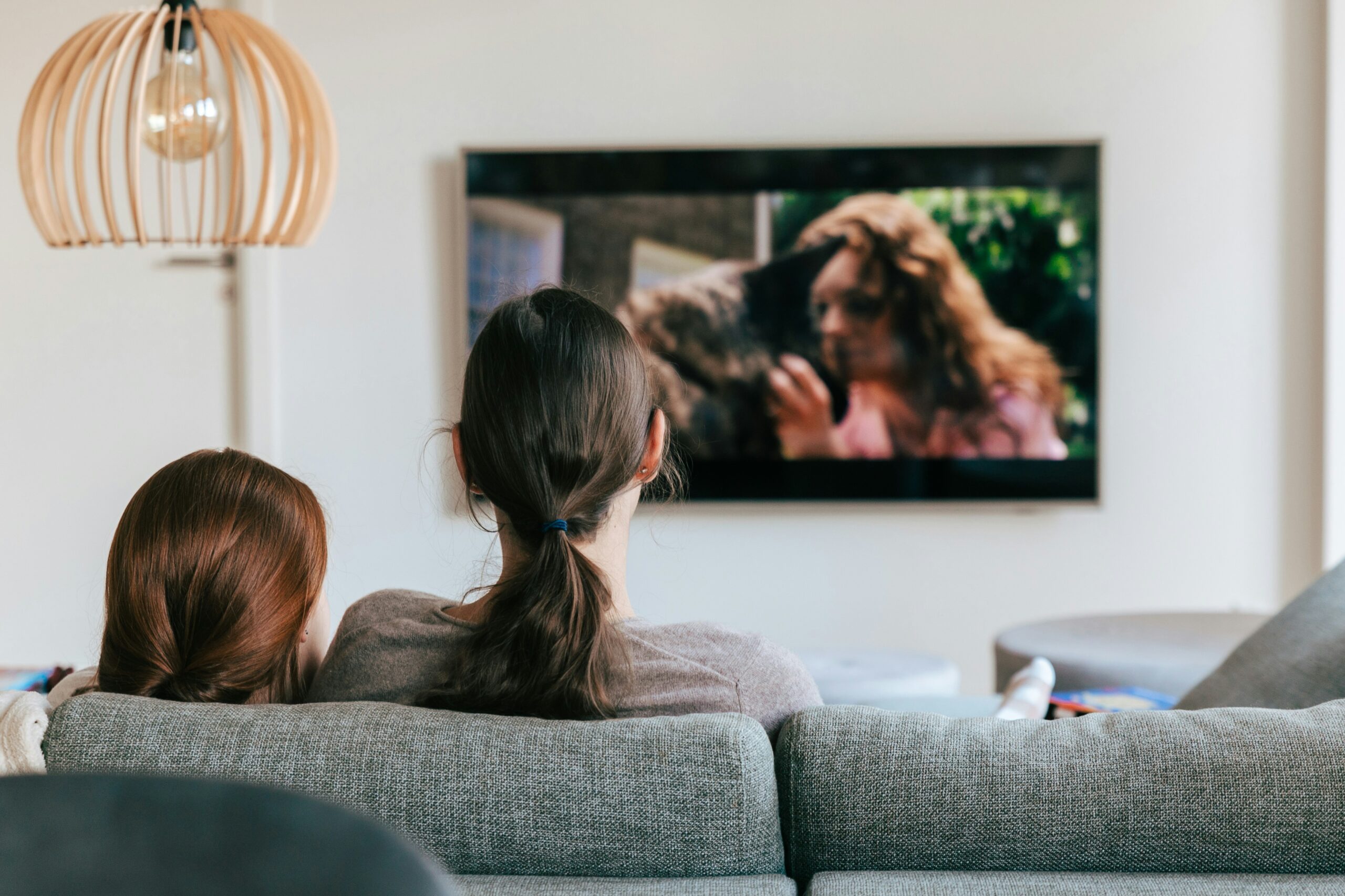Two people sit close on a gray sofa watching a wall-mounted TV showing a couple kissing, with a wooden lampshade to the left in a bright living room.