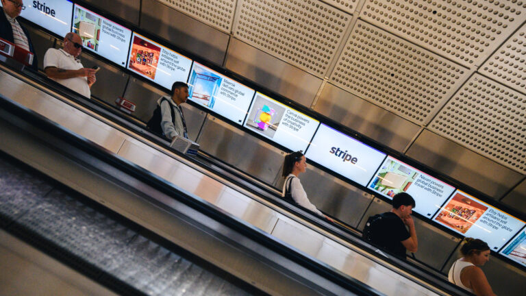 180526 2026 Finance London London Underground Rail Ribbons - Excite Out Of Home - Excite Out Of Home People ride a moving staircase in a modern exhibit with Stripe digital displays along the wall behind them.
