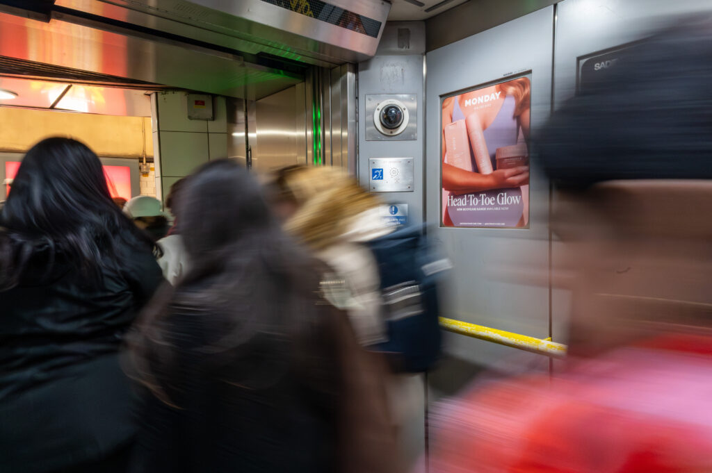 Blurred crowd of passengers in a moving subway car, with a wall-ad featuring MONDAY Head-To-Toe Glow visible on the right and a circular security camera nearby.