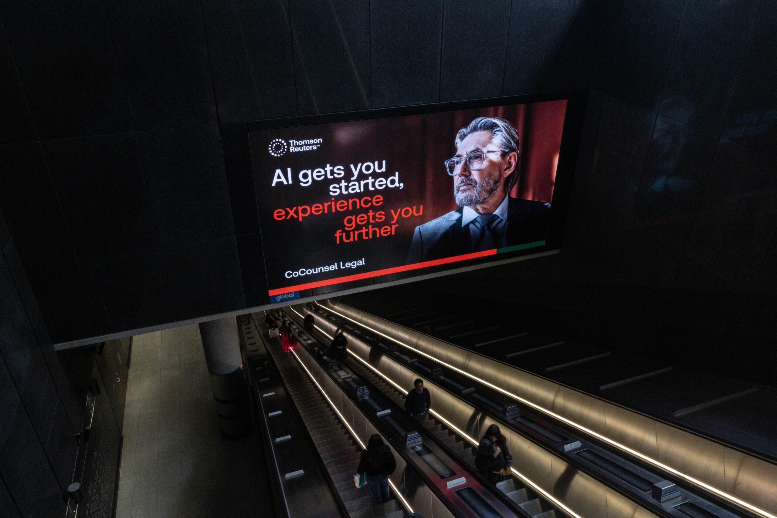 Large digital advertisement in a dark concourse reading 'AI gets you started, experience gets you further' with a suited man on the right and Thomson Reuters logo above.